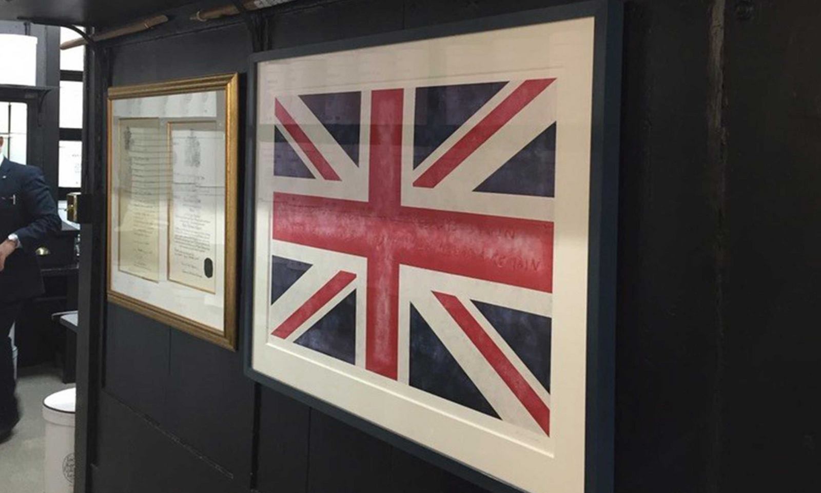 A large framed Union Jack flag print by Martin Wade hangs on a dark wall, with a smaller framed document visible beside it. 