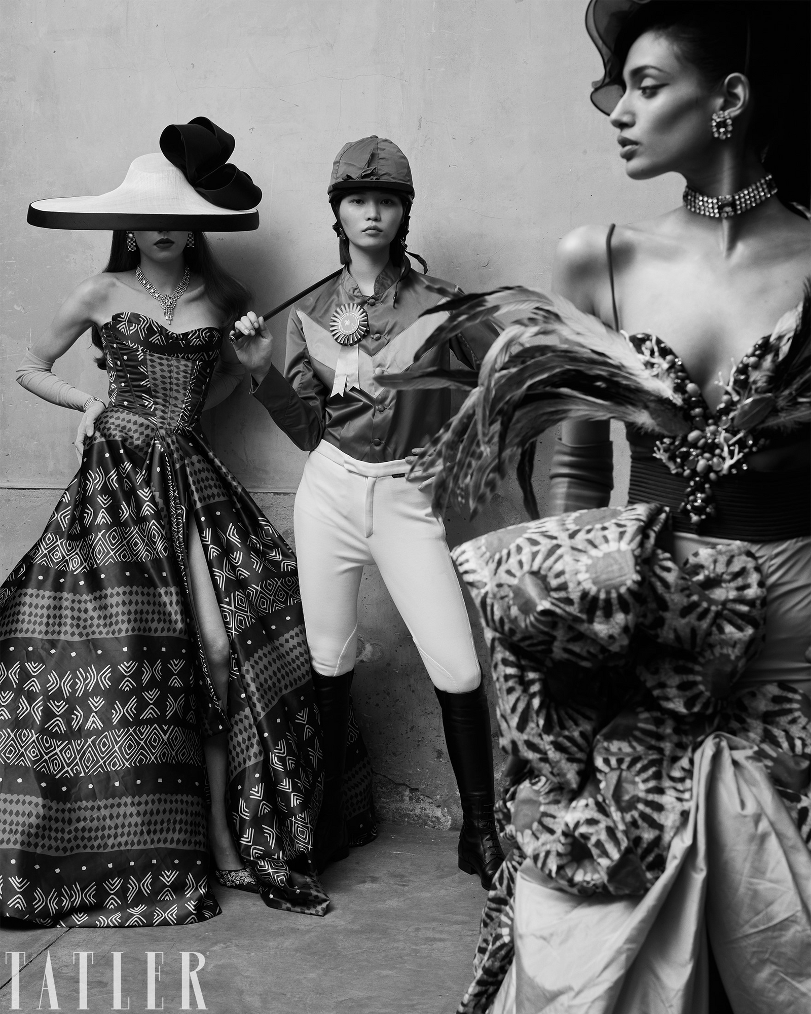 Three fashion models wearing Lock & Co. Hats pose in a black and white editorial photograph against a textured wall, showcasing elegant vintage-inspired fashion looks with patterned gowns, wide-brimmed hats, equestrian attire, and dramatic feathered accessories.